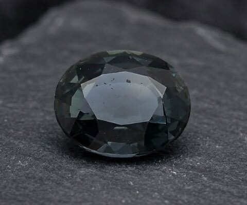 Natural oval-cut teal green blue sapphire gemstone on dark stone surface.