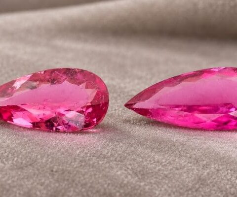 Matching pair of natural bright pink pear-cut tourmaline gemstones on soft beige fabric.