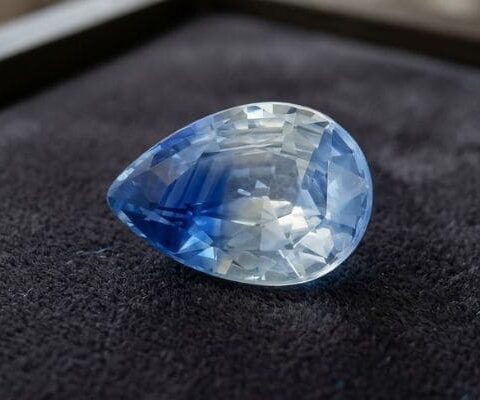 Natural unheated pear-cut bi-color sapphire gemstone displaying blue and white tones on dark velvet fabric.