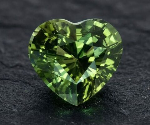 Natural unheated heart-cut blue green sapphire gemstone on dark stone surface.