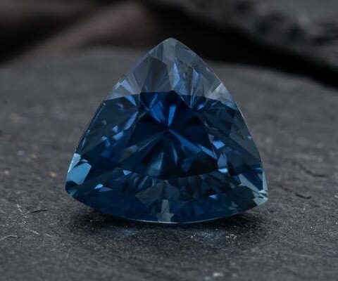 Natural unheated trillion-cut deep blue sapphire gemstone on dark stone surface.