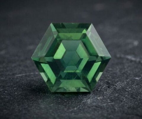 Natural unheated hexagonal-cut bluish green sapphire gemstone on dark stone surface.