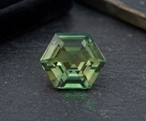 Natural unheated hexagonal-cut bluish green sapphire gemstone on dark stone surface.