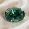 Natural unheated oval-cut greenish blue sapphire gemstone on soft cream fabric.