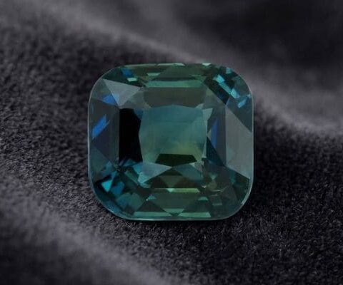 Natural unheated cushion-cut greenish blue sapphire gemstone on dark velvet fabric.