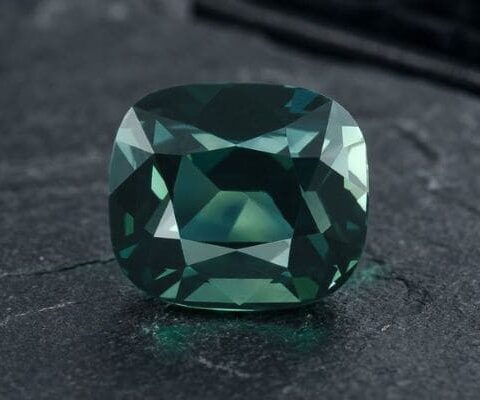 Natural unheated cushion-cut greenish blue sapphire gemstone on dark stone surface.